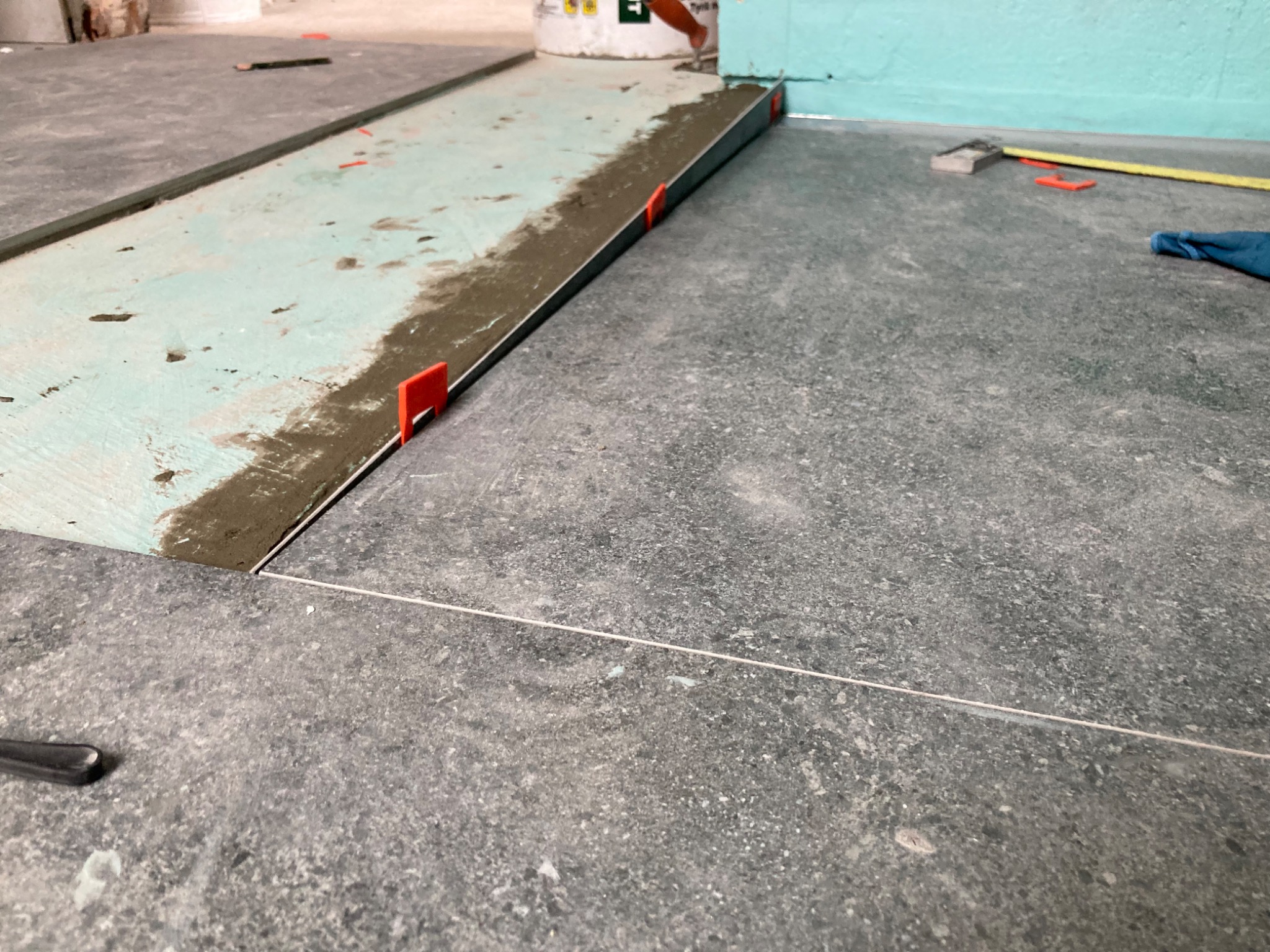 Fragment podłogi w trakcie układania płytek, widoczne płytki, klej, poziomica i sznurek wyznaczający linię, użyte klipsy poziomujące.