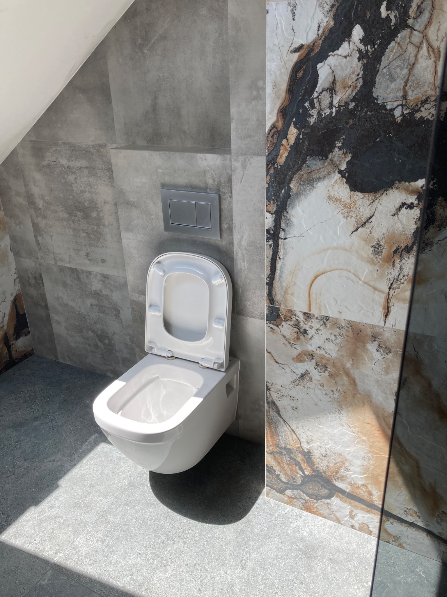 Nowoczesna toaleta podwieszana z otwartą deską, zamontowana w łazience wykończonej szarymi płytkami imitującymi beton i płytkami z wzorem marmuru z brązowymi żyłami, oświetlona naturalnym światłem...