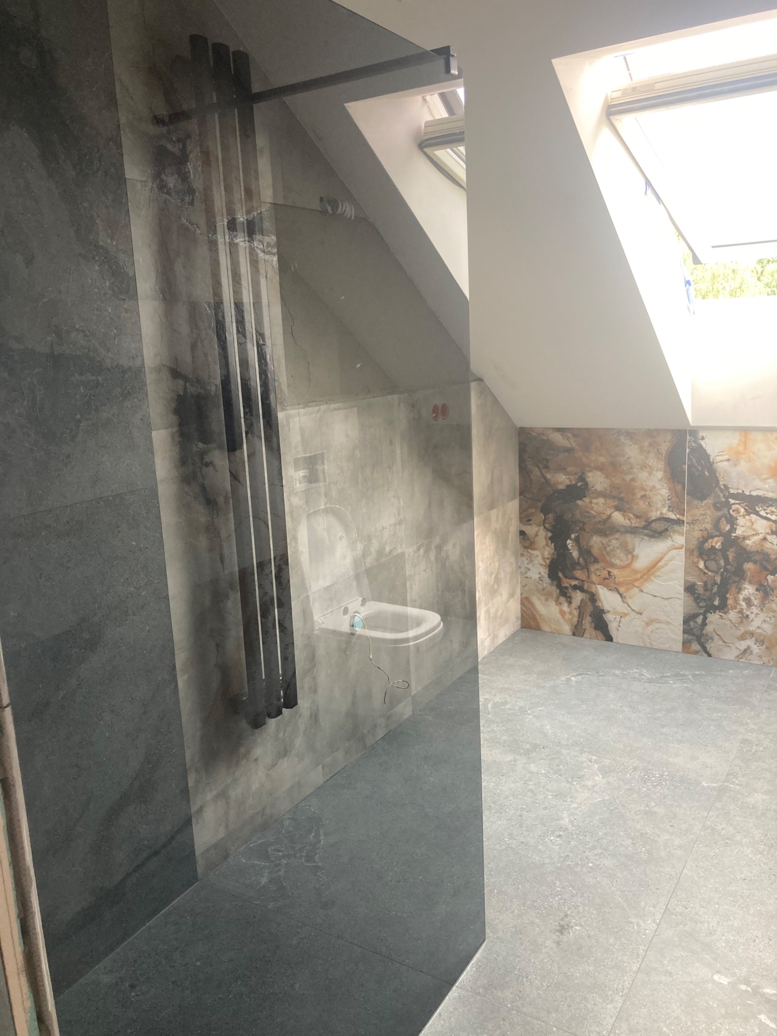 Wnętrze łazienki na poddaszu z oknem dachowym, wykończonej szarymi i brązowymi płytkami imitującymi kamień, z widocznym grzejnikiem drabinkowym i częściowo widoczną toaletą.