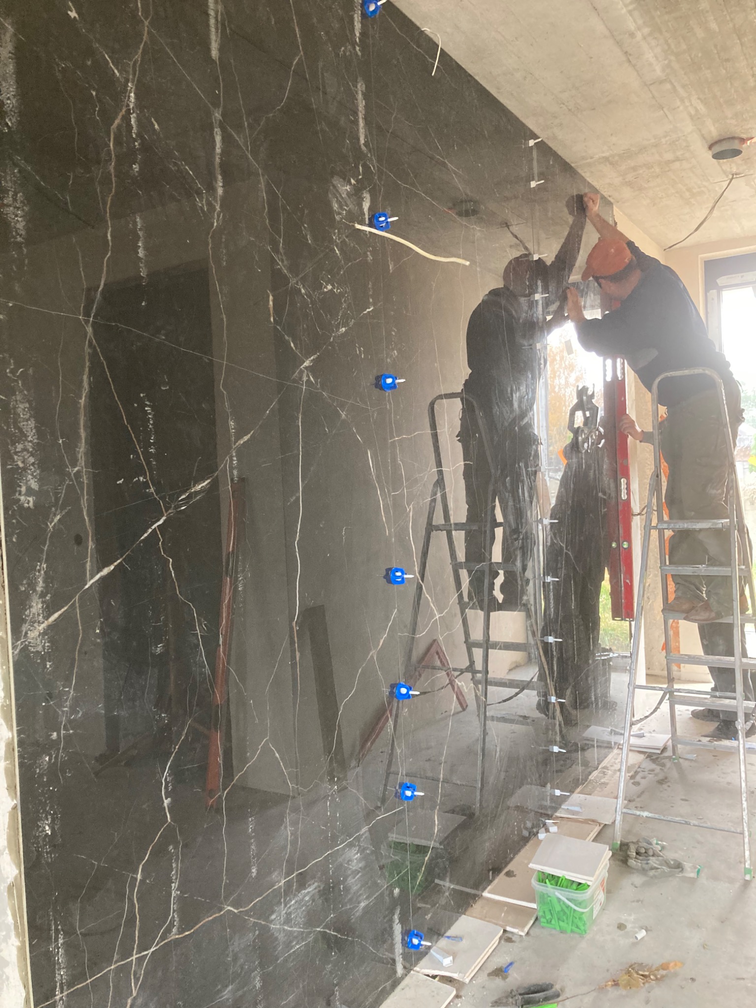 Montaż czarnych, marmurkowych płyt na ścianie, z użyciem poziomicy i krzyżyków dystansowych, przez dwóch pracowników na drabinach.