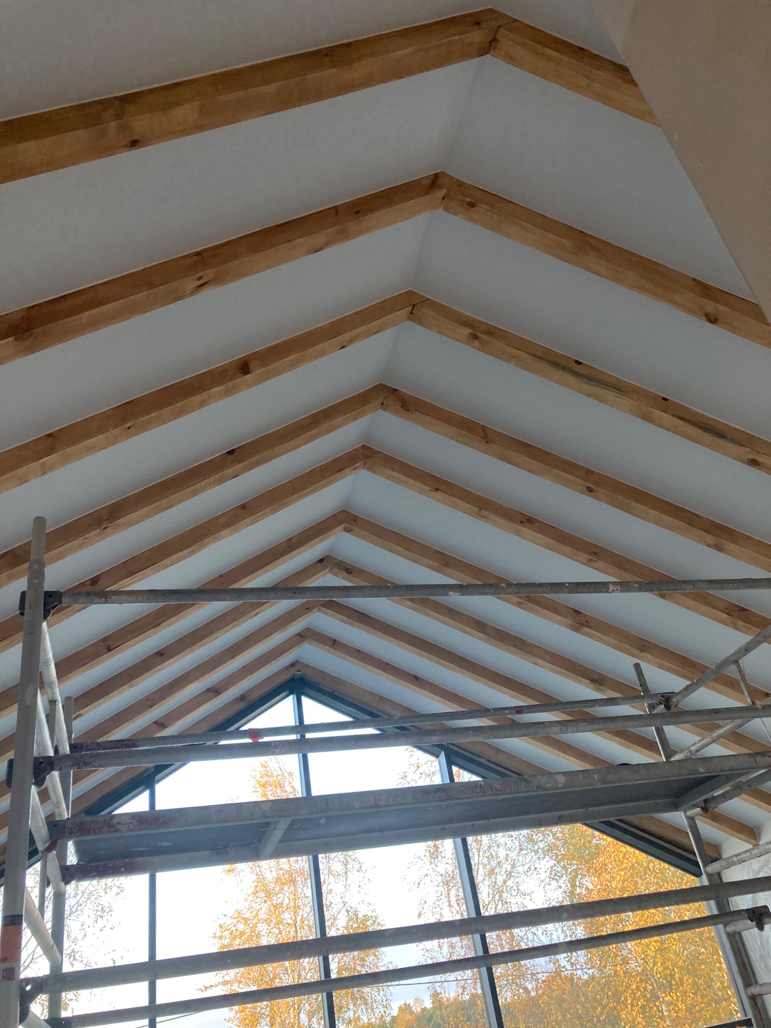 Wnętrze w trakcie wykańczania: widok na drewnianą konstrukcję dachu z izolacją, uchwycony ze stojącego rusztowania, z widokiem na jesienne drzewa za oknem.