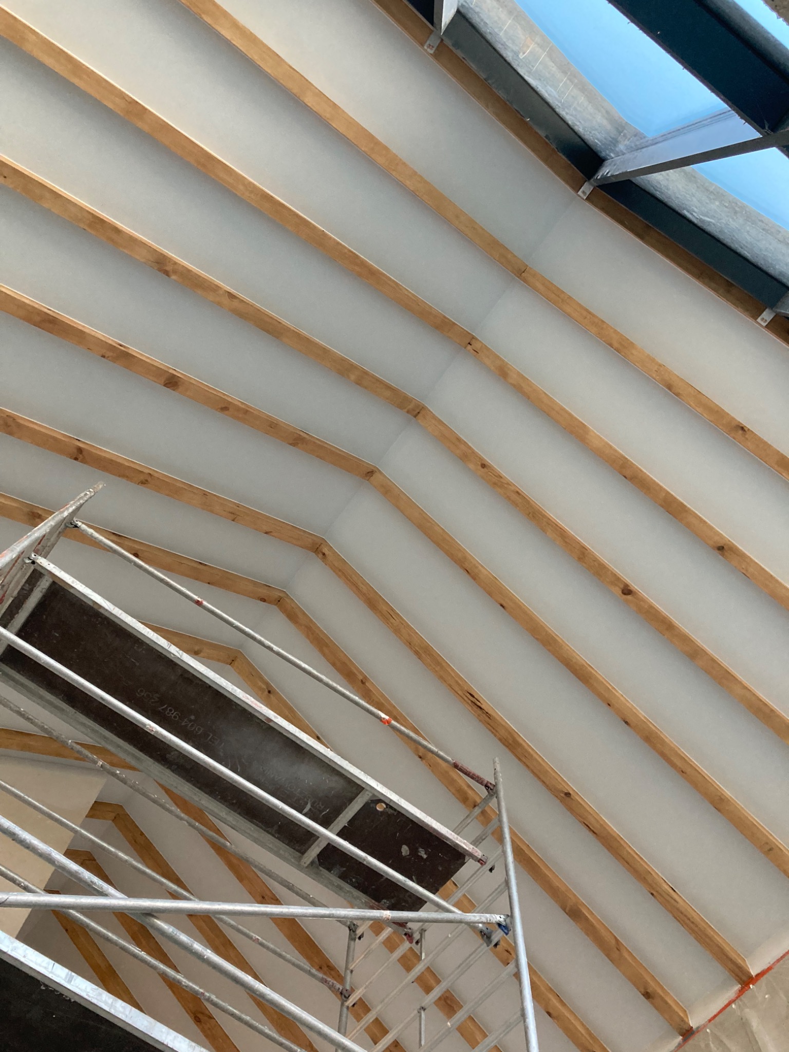 Sufit poddasza z widocznymi drewnianymi belkami i białą powierzchnią między nimi, częściowo zasłonięty przez rusztowanie.