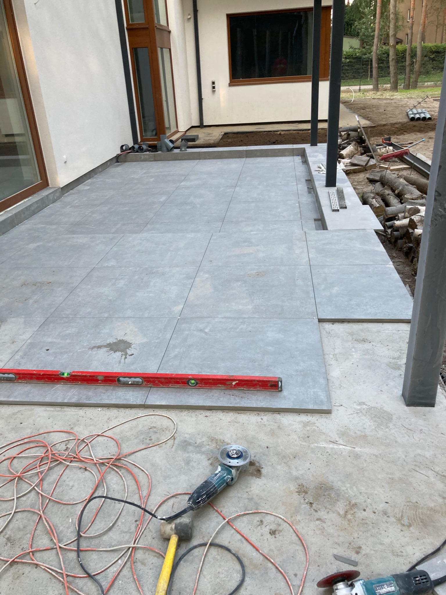 Układanie szarych płytek na tarasie domu jednorodzinnego, widoczny poziomica, szlifierka kątowa i przewody elektryczne.