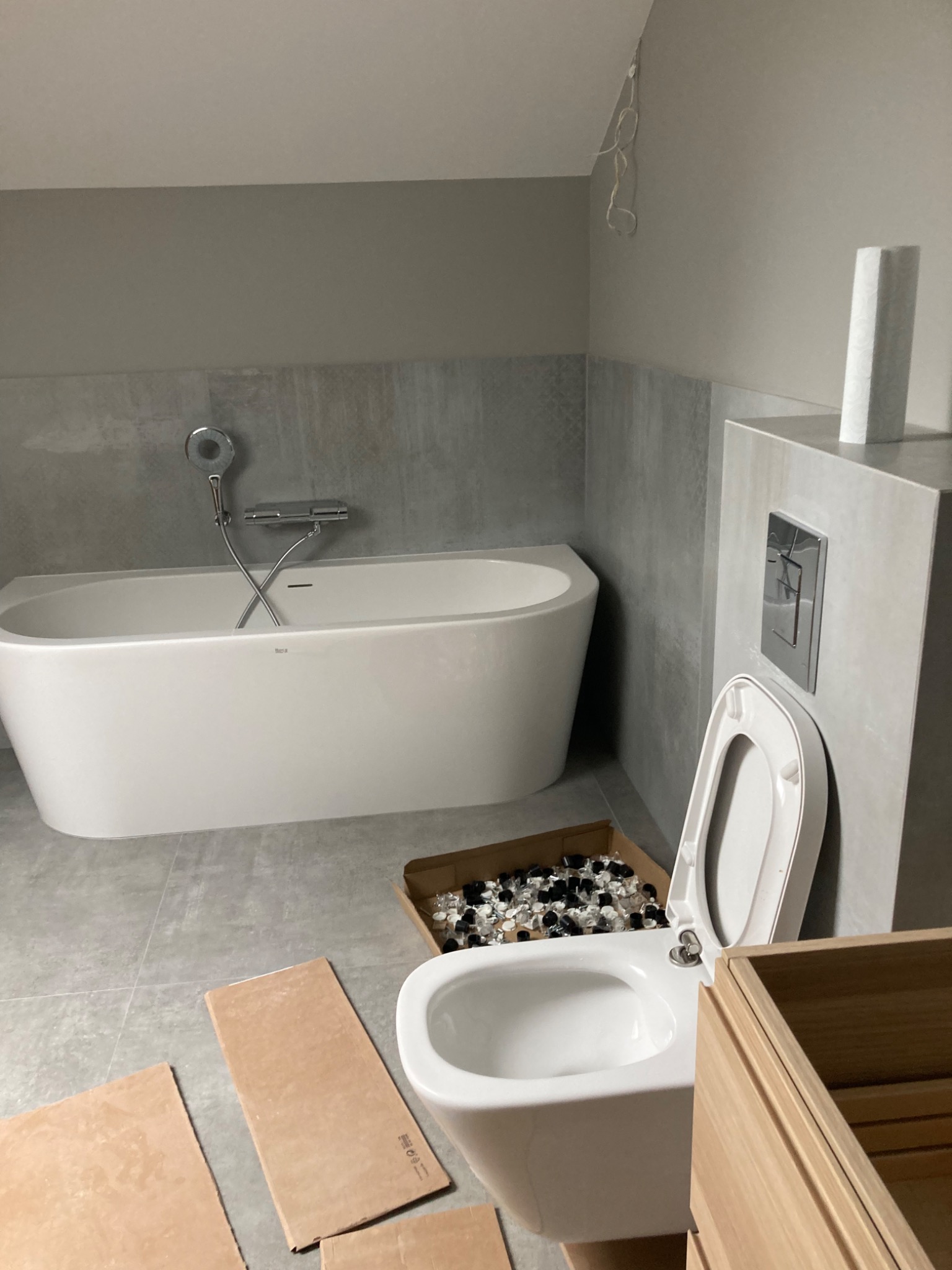 Nowoczesna łazienka w trakcie montażu: biała wanna wolnostojąca, podwieszana toaleta z otwartą klapą, karton z elementami montażowymi i fragment szafki z jasnego drewna na tle szarych płytek.