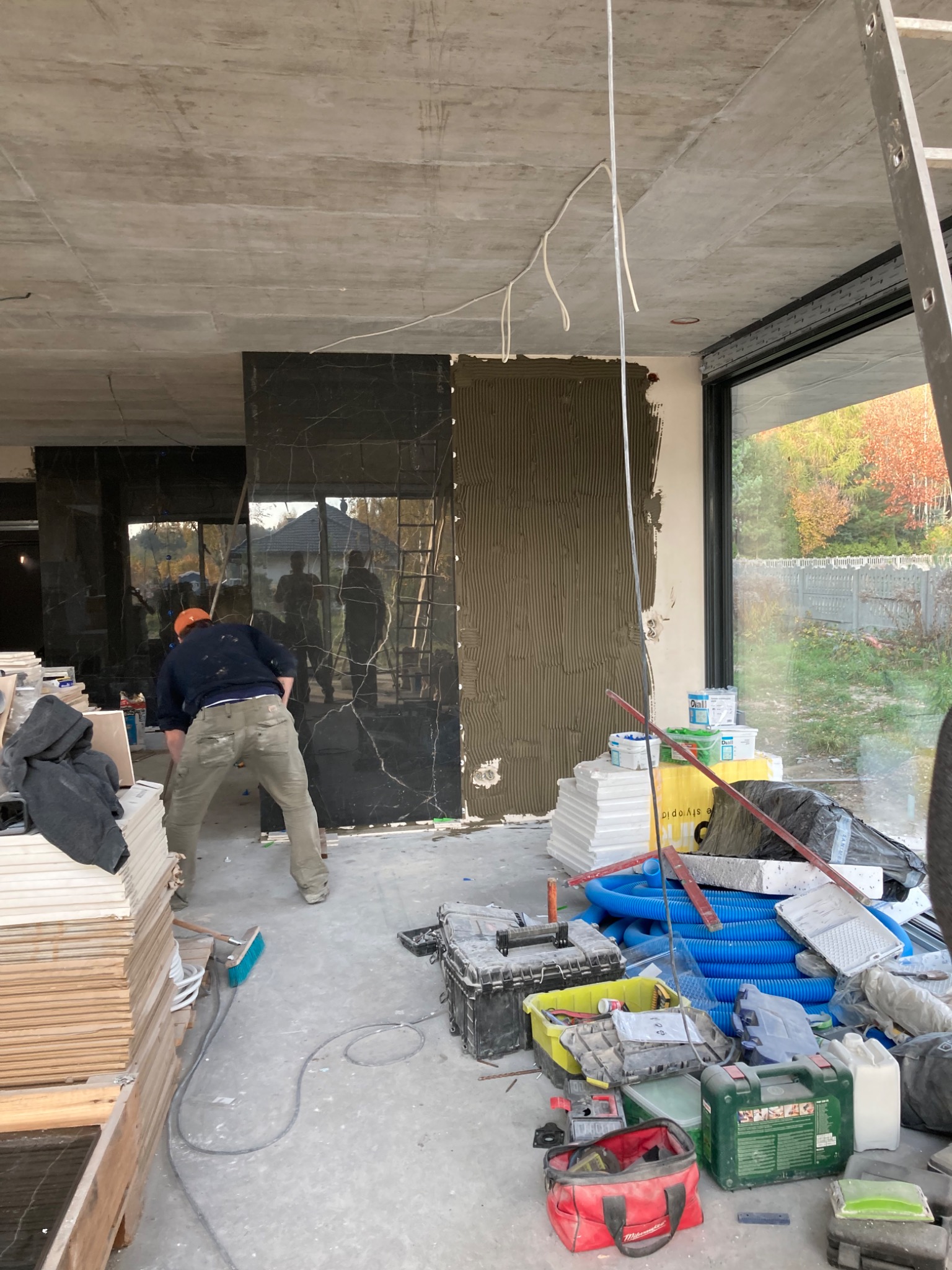 Prace wykończeniowe w surowym wnętrzu: glazurnik w pomarańczowej czapce nakłada klej na ścianę pod czarne płytki imitujące marmur, widoczny stos płytek, narzędzia i materiały budowlane.