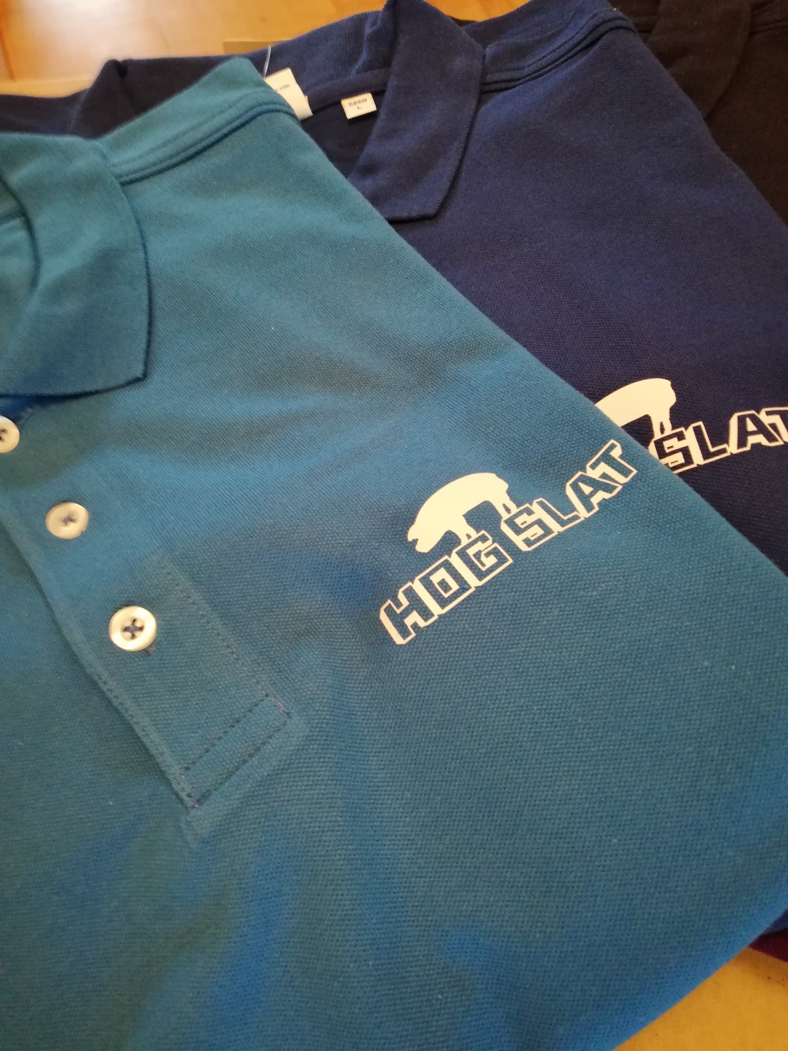 Dwa polo: turkusowe i granatowe z białym nadrukiem logo 'Hog Slat', widoczne guziki i faktura materiału.