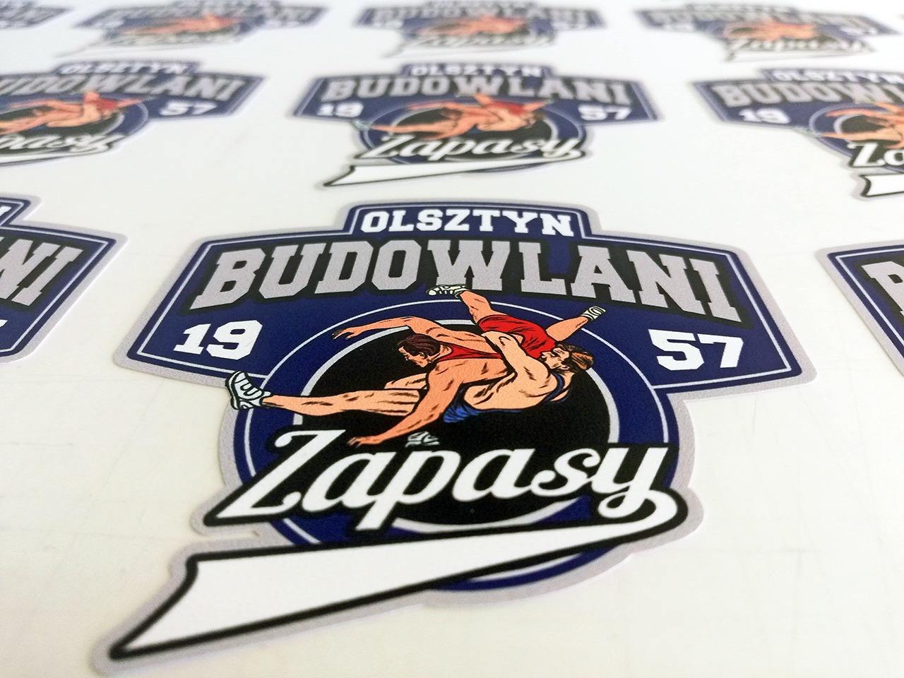 Arkusz naklejek przedstawiających logo klubu zapaśniczego Budowlani Olsztyn z motywem dwóch zapaśników w walce, na białym tle.