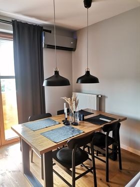 Klimatyzator ścienny zamontowany nad oknem w nowoczesnej jadalni z drewnianym stołem i czarnymi lampami wiszącymi.