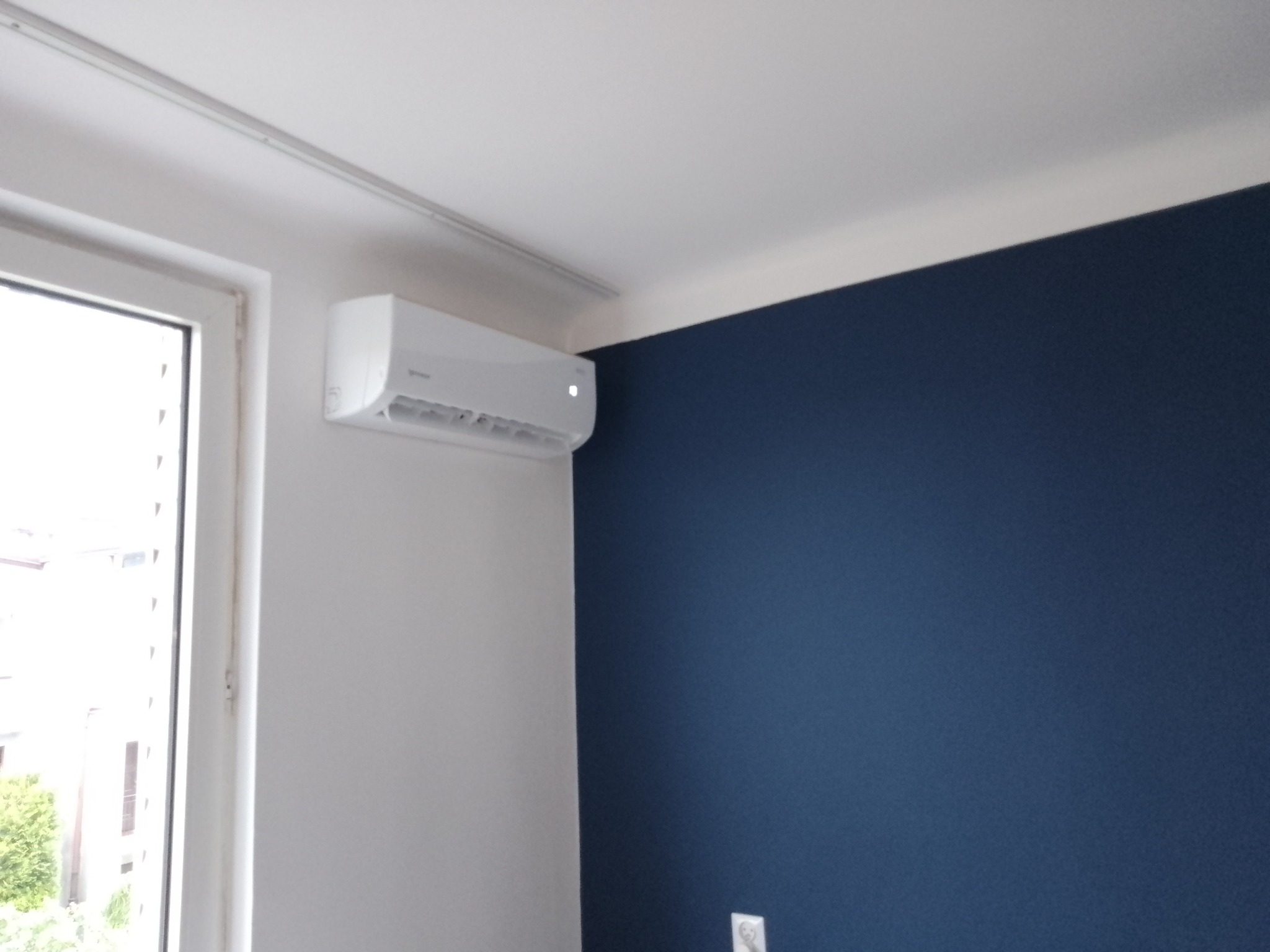 Biały klimatyzator marki Gorenje zamontowany na białej ścianie w pomieszczeniu z oknem i granatową ścianą akcentującą.