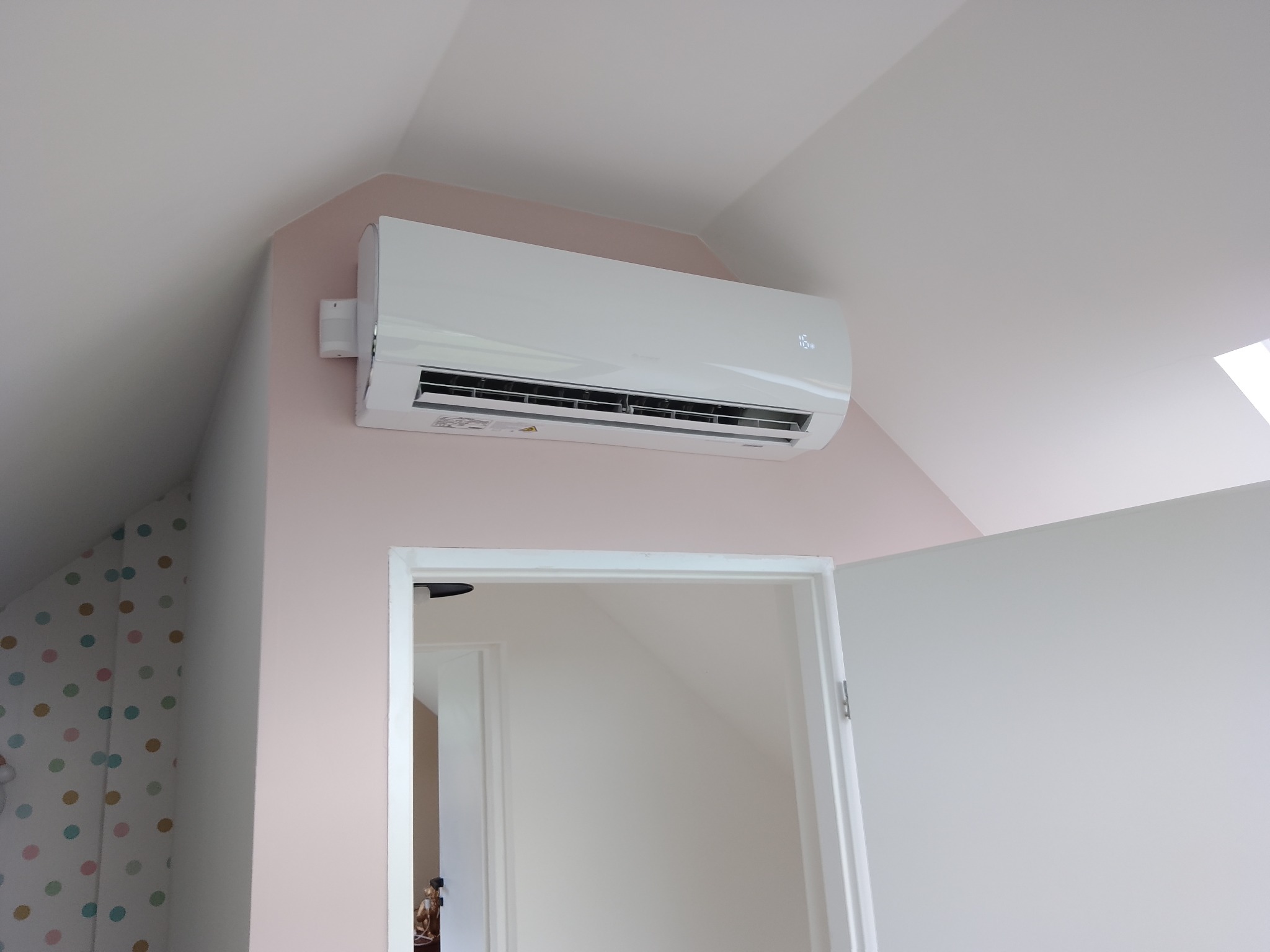 Biały klimatyzator ścienny zamontowany w pokoju na poddaszu, widok z dołu na urządzenie i otwarte drzwi do sąsiedniego pomieszczenia.
