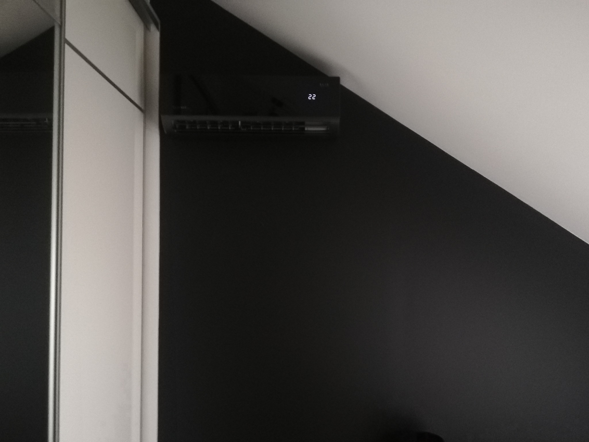 Czarny klimatyzator ścienny zamontowany na skośnej czarnej ścianie w pokoju, obok biała szafa z lustrzanymi drzwiami, wyświetlacz pokazuje temperaturę 22 stopnie.