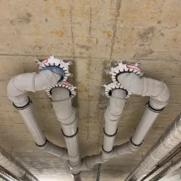 Widok z dołu na szare rury kanalizacyjne przymocowane do surowego betonowego sufitu za pomocą białych kołnierzy przeciwpożarowych i czarnych obejm, dodatkowo widoczne ocieplone rury.