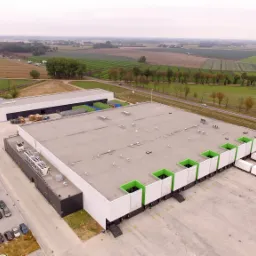 Widok z lotu ptaka na duży budynek magazynowy z płaskim dachem, rampami załadunkowymi z zielonymi akcentami i zaparkowanymi ciężarówkami, otoczony polami i drogą.