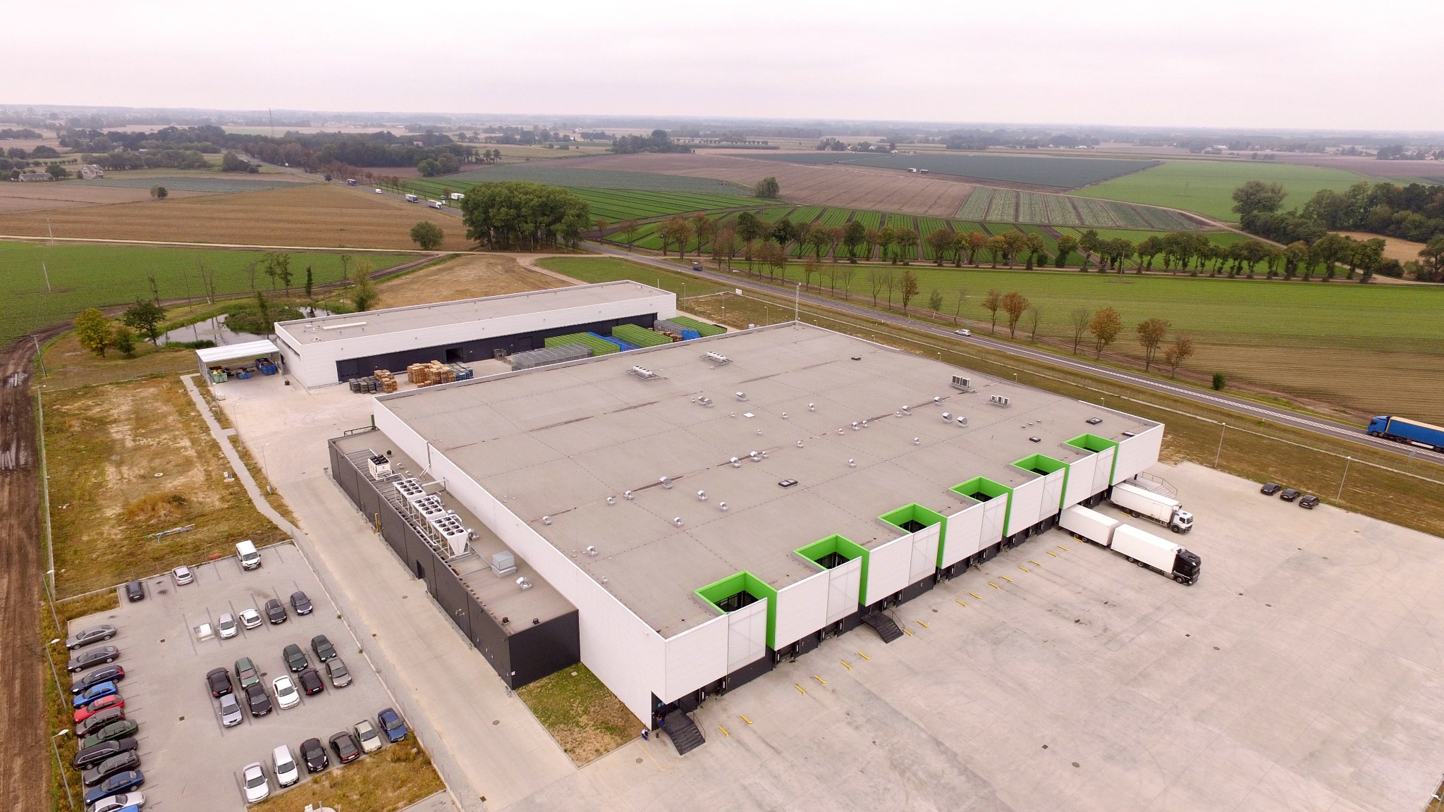 Widok z lotu ptaka na duży budynek magazynowy z płaskim dachem, rampami załadunkowymi z zielonymi akcentami i zaparkowanymi ciężarówkami, otoczony polami i drogą.