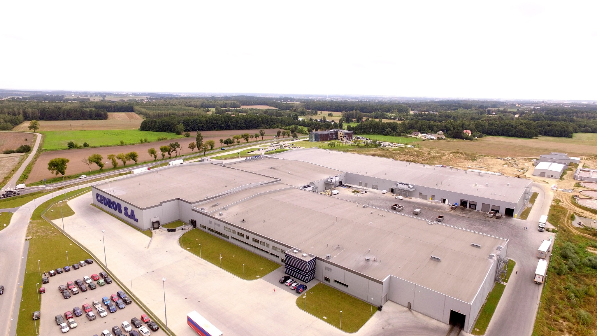 Rozległy kompleks budynków przemysłowych CEDROB S.A. z płaskimi dachami, widok z lotu ptaka, z parkingiem pełnym samochodów i zielonym otoczeniem.