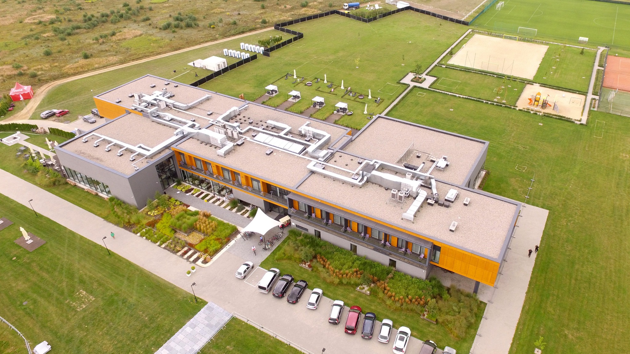 Widok z drona na duży, nowoczesny budynek z płaskim dachem, systemami wentylacyjnymi, pomalowanymi na pomarańczowo fragmentami elewacji i zadbanym otoczeniem z parkingiem, zielenią i elementami...