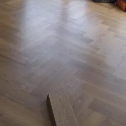 Ułożona podłoga z paneli w jodełkę, widoczne narzędzia w tle, niedokończony fragment z pojedynczym panelem na pierwszym planie.