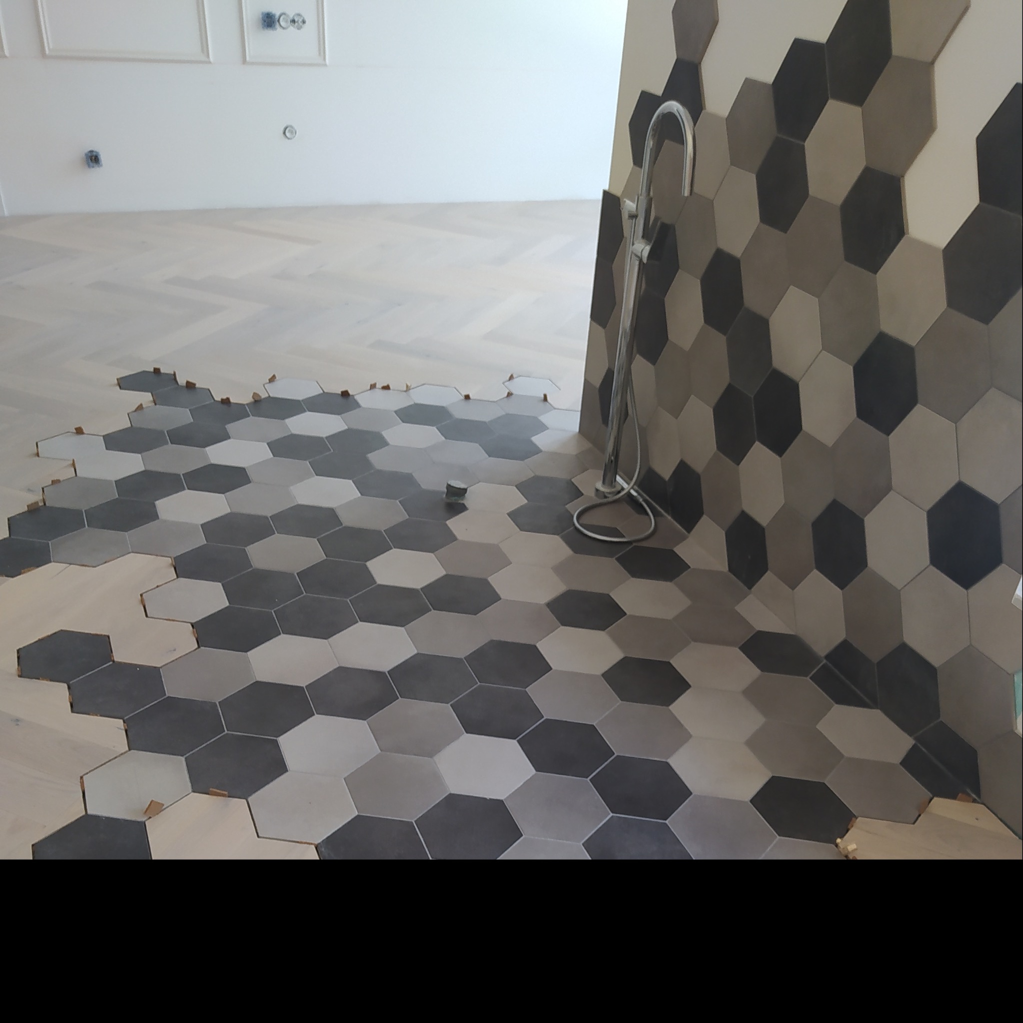 Układanie heksagonalnych płytek w odcieniach szarości na podłodze i ścianie łazienki, widoczne drewniane kliny dystansowe i fragment drewnianej podłogi w jodełkę.