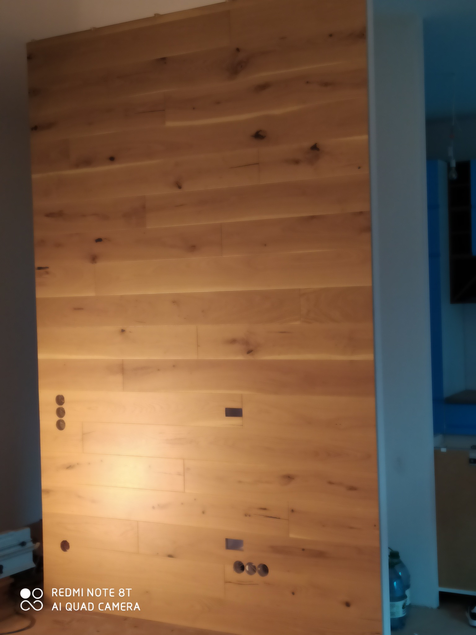 Ściana wykończona panelami drewnianymi w ciepłym odcieniu, z widocznymi otworami na gniazdka elektryczne i fragmentem niebieskiego pomieszczenia w tle.