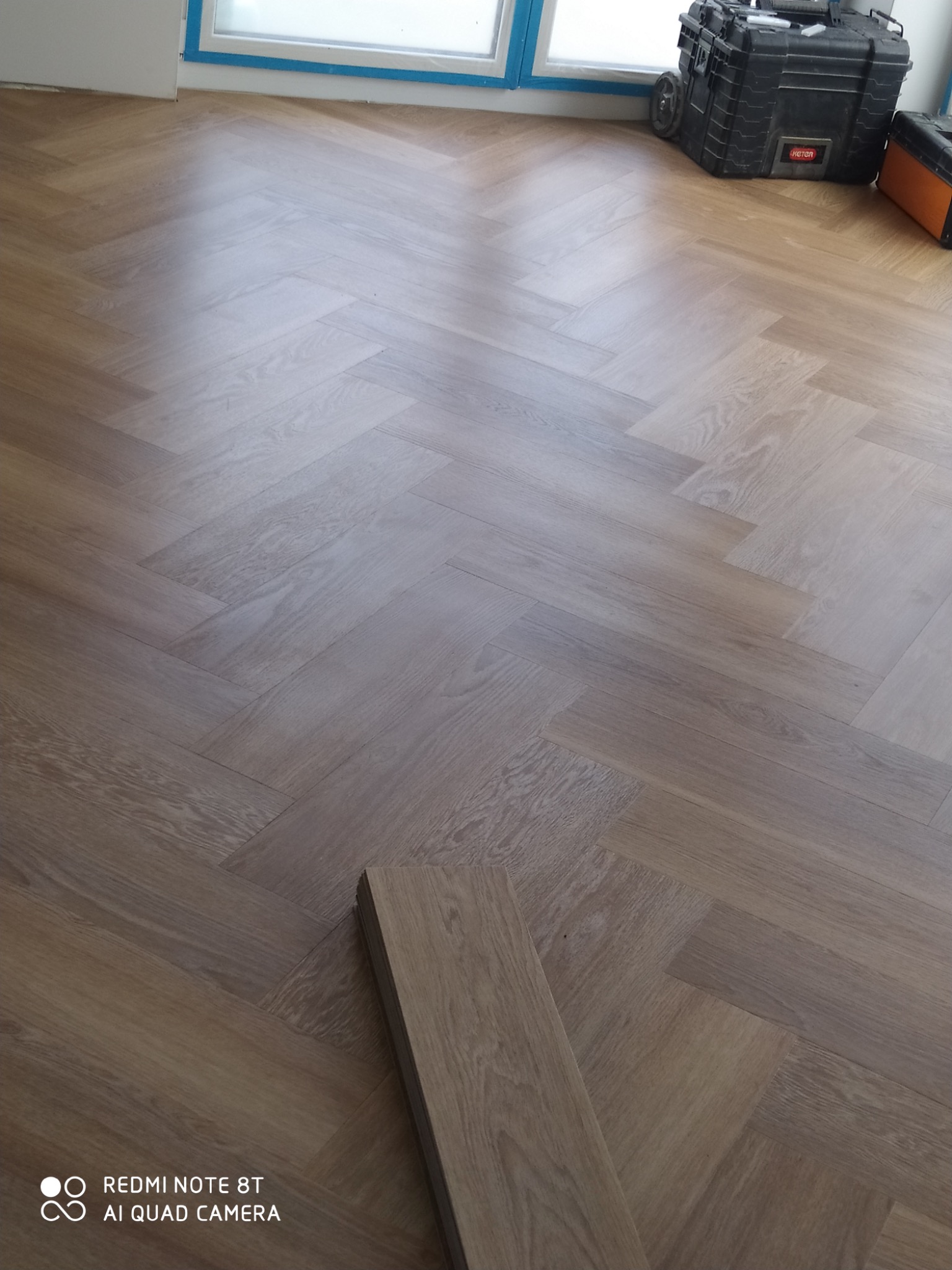 Ułożona podłoga z paneli w jodełkę, widoczne narzędzia w tle, niedokończony fragment z pojedynczym panelem na pierwszym planie.