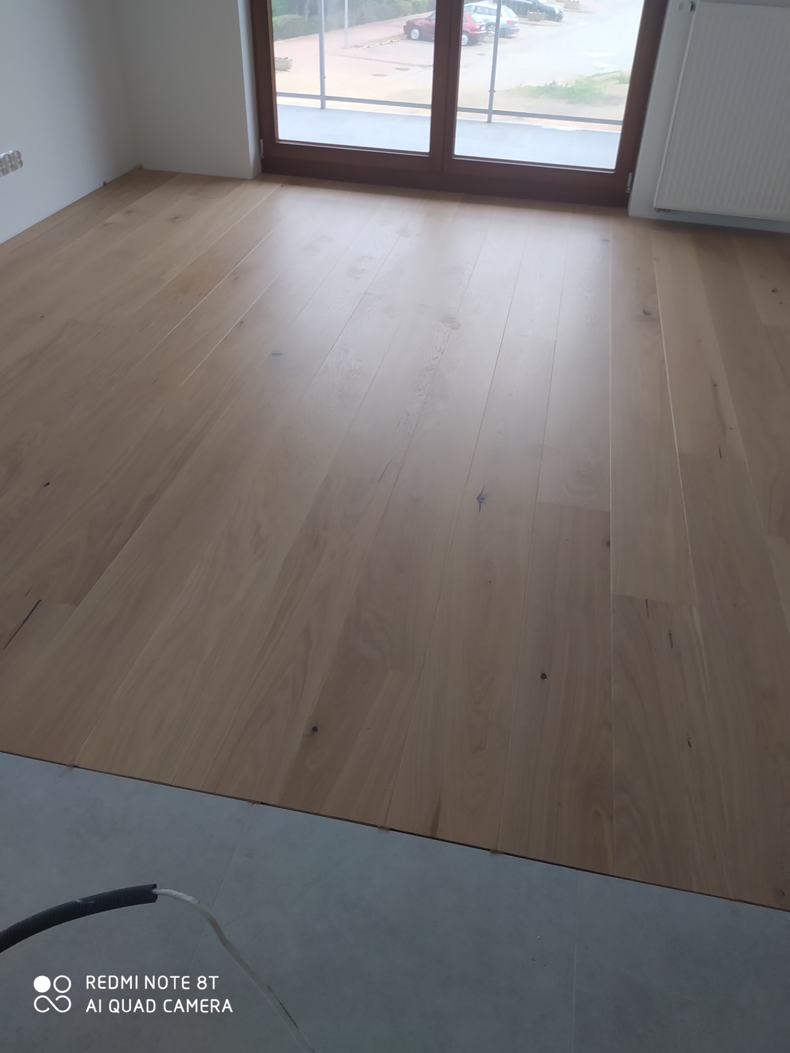 Jasne, lakierowane deski parkietowe ułożone w nowym pomieszczeniu, widok z niskiego kąta, z widocznym fragmentem grzejnika i okna balkonowego w tle.