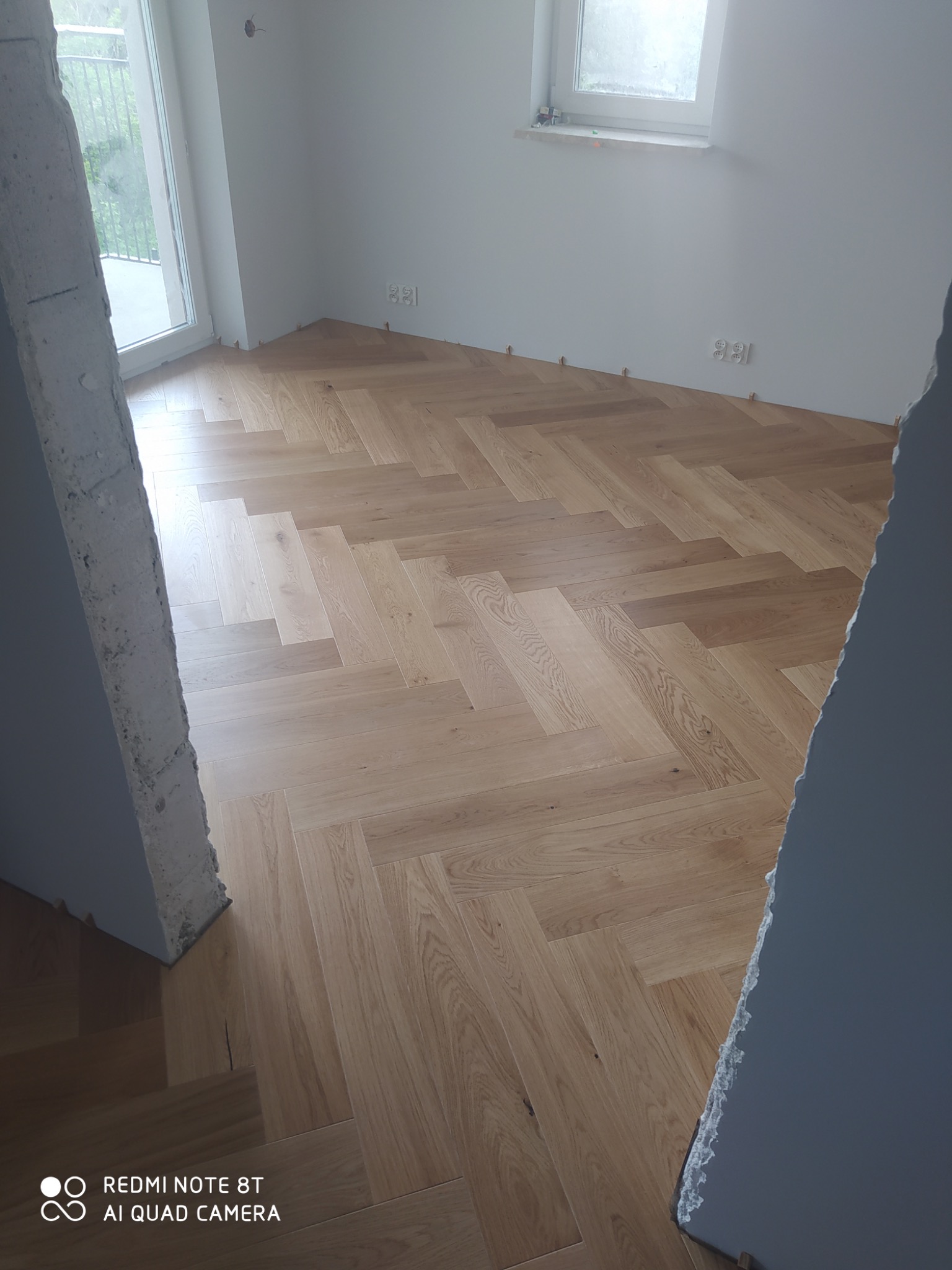 Jasny parkiet ułożony we wzór jodełki francuskiej, widoczne kliny dystansowe przy ścianach, surowe ściany w trakcie wykończenia, widok z sąsiedniego pomieszczenia.