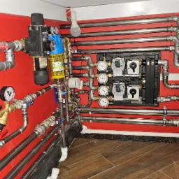 Złożona instalacja hydrauliczna z licznymi rurami, zaworami, manometrami i pompami obiegowymi na czerwonej ścianie, widoczne elementy systemu grzewczego i wodnego w pomieszczeniu.