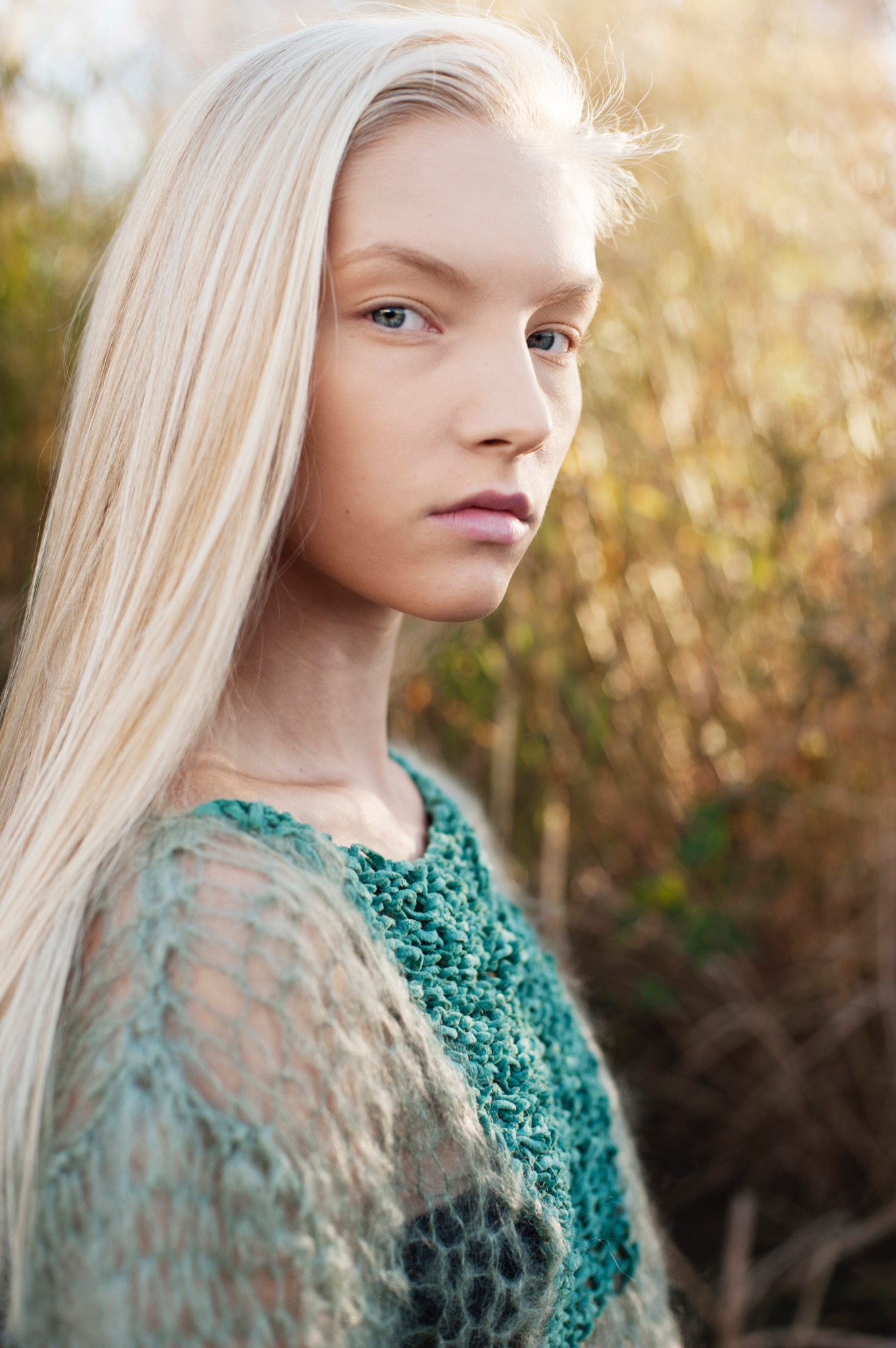 Portret kobiety o jasnych włosach i niebieskich oczach, ubrana w ażurowy, turkusowy sweter na tle rozmytych, jesiennych traw.