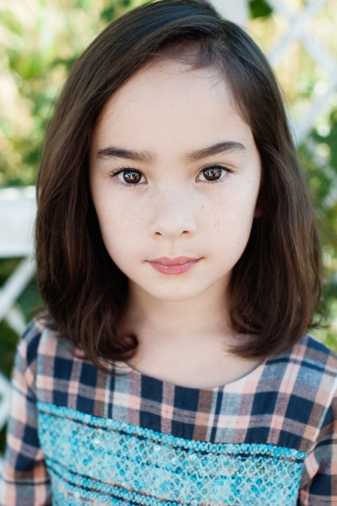 Portret młodej dziewczynki z brązowymi włosami i piegami, ubrana w bluzkę w kratę z turkusowym zdobieniem, w naturalnym świetle.
