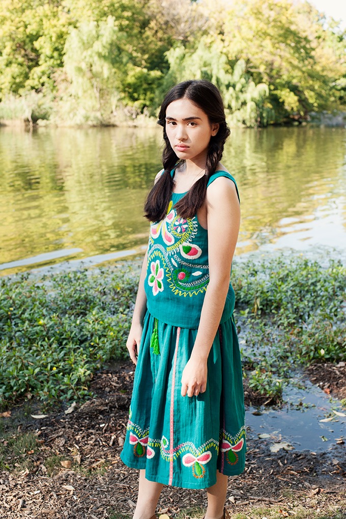 Młoda kobieta w turkusowym, haftowanym komplecie (bluzka i spódnica) pozuje na tle jeziora i zieleni, z włosami zaplecionymi w dwa warkocze.