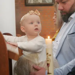 Dziewczynka z kokardą na głowie, opierająca się o drewniany stół, obok świeca z symbolami religijnymi, w tle mężczyzna w jasnej marynarce.