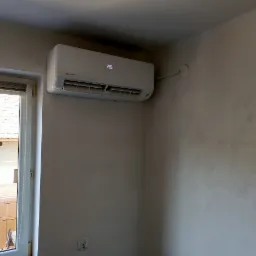 Biały klimatyzator ścienny marki Rotenso zamontowany w pokoju z dekoracyjną, szarą ścianą, widoczny fragment okna z roletą i drabina na zewnątrz.