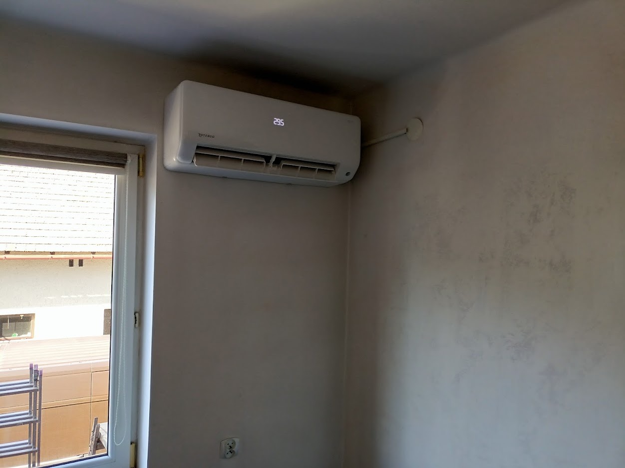 Biały klimatyzator ścienny marki Rotenso zamontowany w pokoju z dekoracyjną, szarą ścianą, widoczny fragment okna z roletą i drabina na zewnątrz.