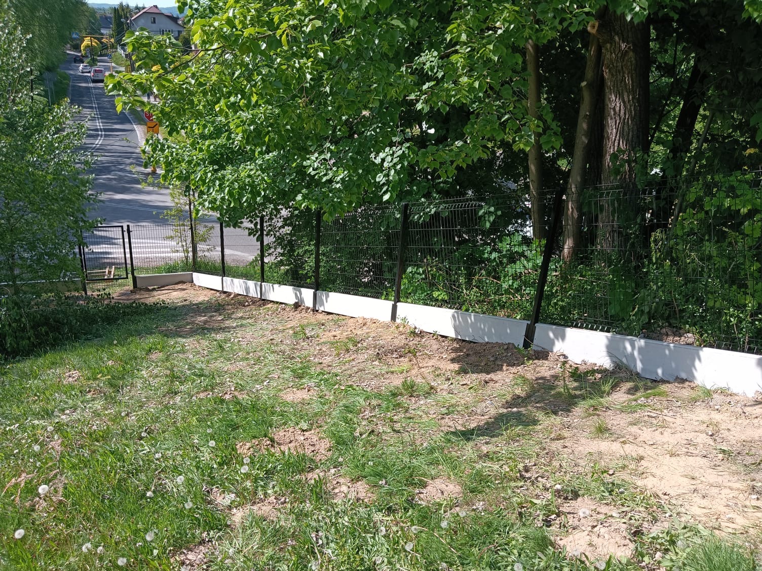 Nowoczesne ogrodzenie panelowe z podmurówką betonową na nierównym terenie, widoczne od strony posesji, z perspektywą na ulicę i domy w tle.