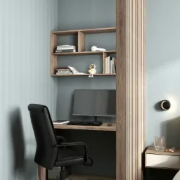 Kompaktowe biuro domowe z biurkiem wbudowanym w regał, czarne krzesło obrotowe, półki z książkami i figurką astronauty, oddzielone od sypialni drewnianą ścianką z listew.