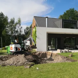 Mini koparka Bobcat E19 wykonuje wykop pod fundamenty domu z nowoczesną architekturą, widoczny fragment tarasu i meble ogrodowe.