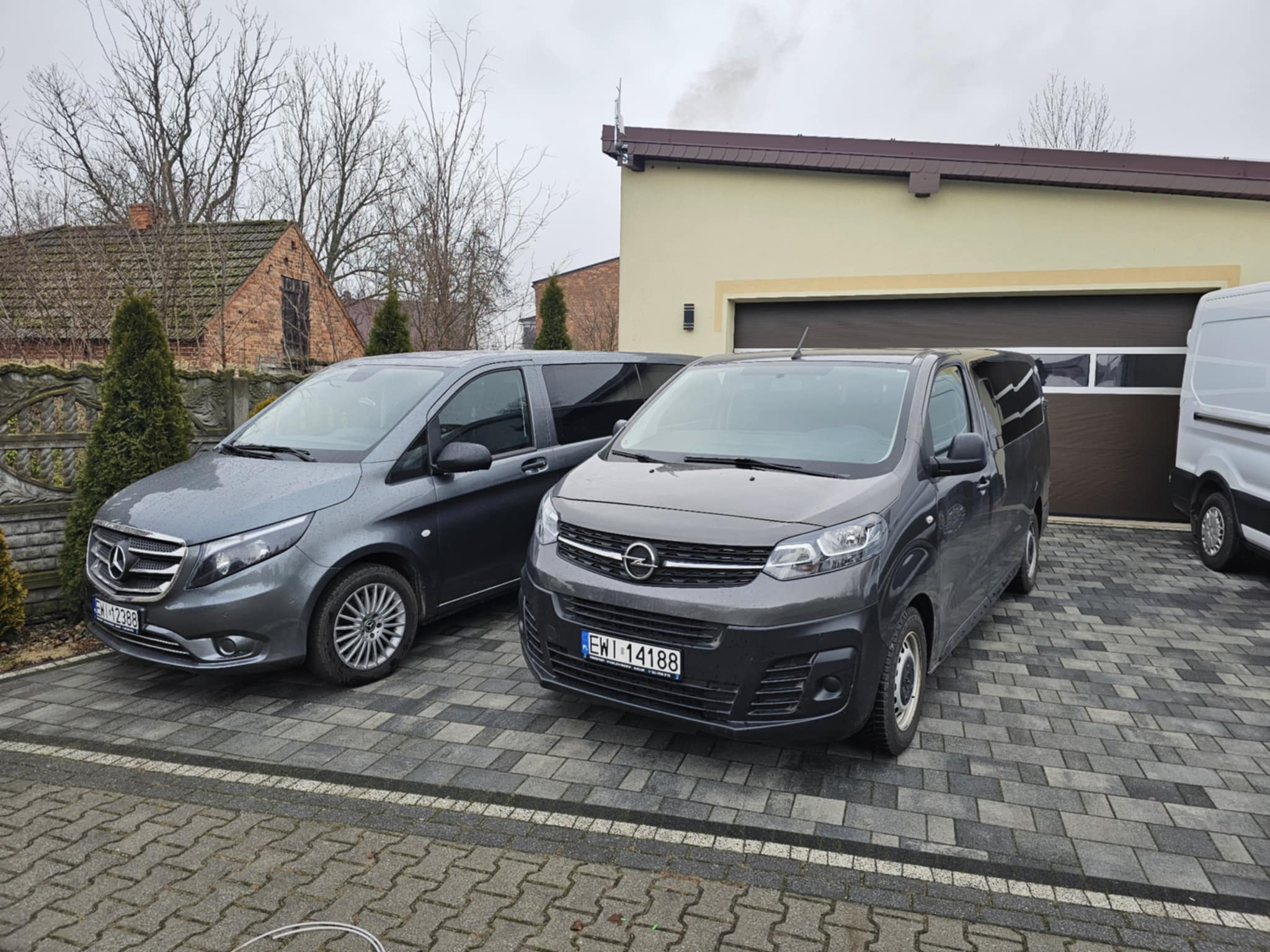 Dwa szare busy marki Mercedes Vito i Opel Vivaro zaparkowane na kostce brukowej przed budynkiem z brązową bramą garażową, w tle widoczny budynek z cegły i komin, pochmurne niebo.