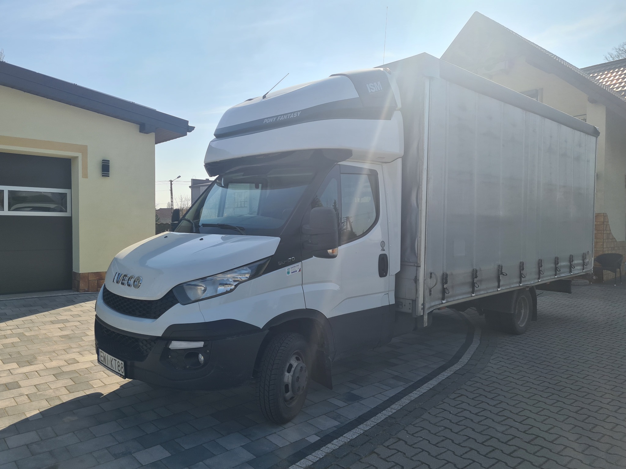 Biały samochód dostawczy Iveco z zabudową kontenerową i plandeką, zaparkowany na kostce brukowej przed budynkiem w słoneczny dzień.