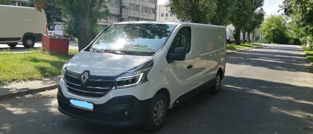 Biały samochód dostawczy Renault Trafic zaparkowany na ulicy w słoneczny dzień, z widocznym budynkiem w tle.