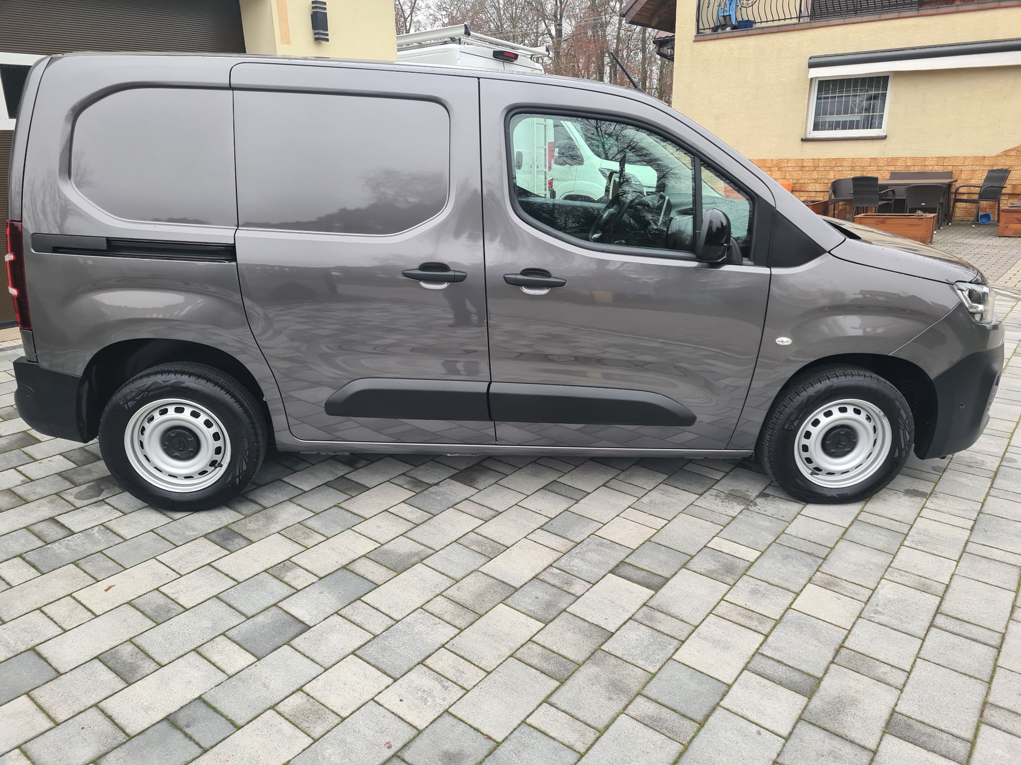 Szary samochód dostawczy typu furgon z przesuwnymi drzwiami bocznymi, zaparkowany na szarej kostce brukowej przed budynkiem z beżową elewacją.