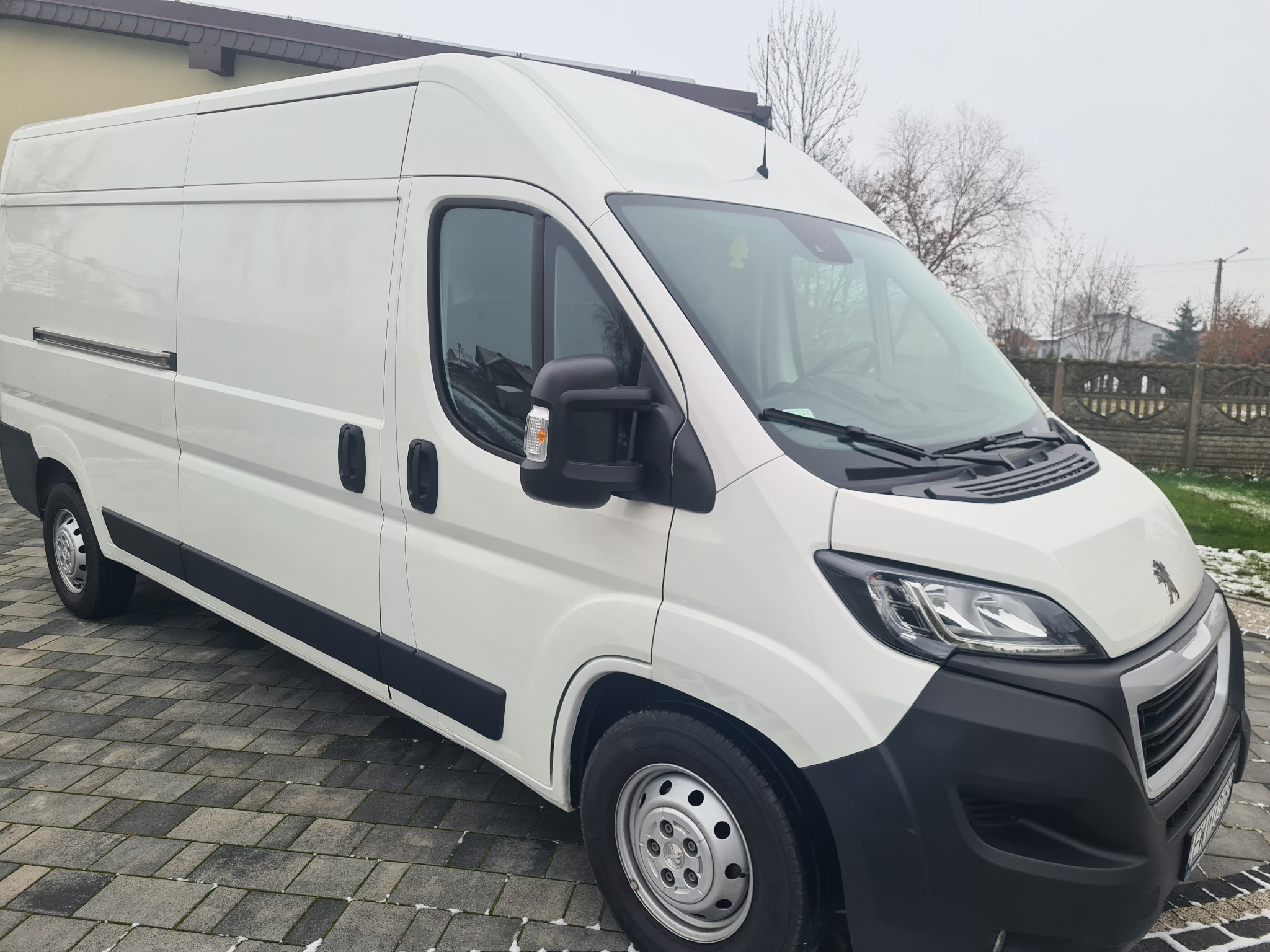 Biały samochód dostawczy Peugeot Boxer zaparkowany na szarej kostce brukowej, widok z przodu i z boku, pochmurny dzień.