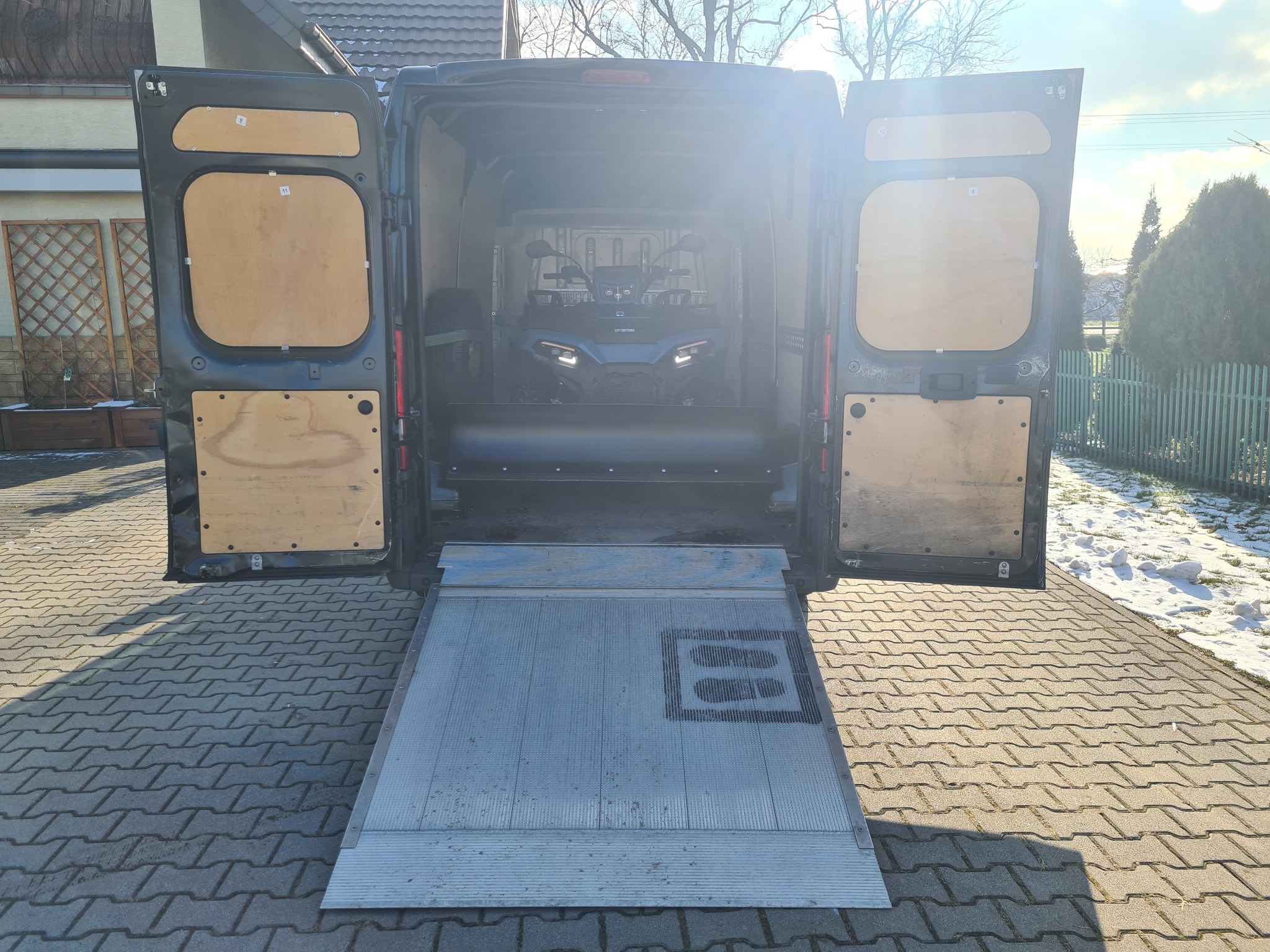 Czarny bus z otwartymi drzwiami i rampą załadunkową, wewnątrz quad ATV, widoczne ślady stóp na rampie.