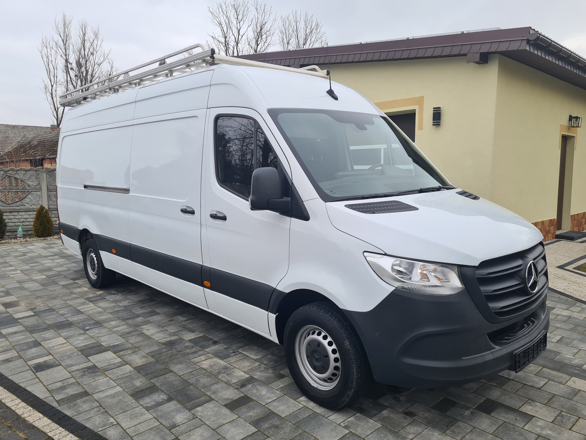 Biały samochód dostawczy Mercedes-Benz Sprinter z relingami dachowymi, zaparkowany na szarej kostce brukowej przed budynkiem z beżową elewacją i brązowym dachem.