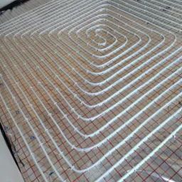 Białe rury ogrzewania podłogowego ułożone spiralnie na folii z czerwoną siatką, widoczne niebieskie klipsy mocujące.