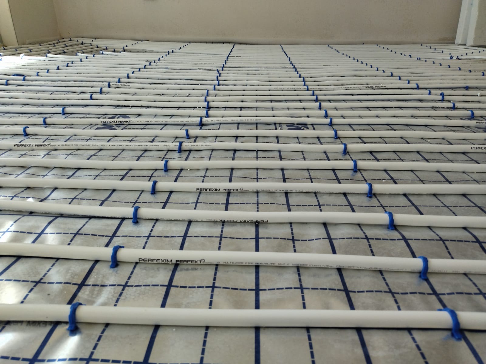 Białe rury ogrzewania podłogowego, mocowane niebieskimi klipsami do folii z nadrukowaną siatką, ułożone równolegle na podłożu w jasnym pomieszczeniu.