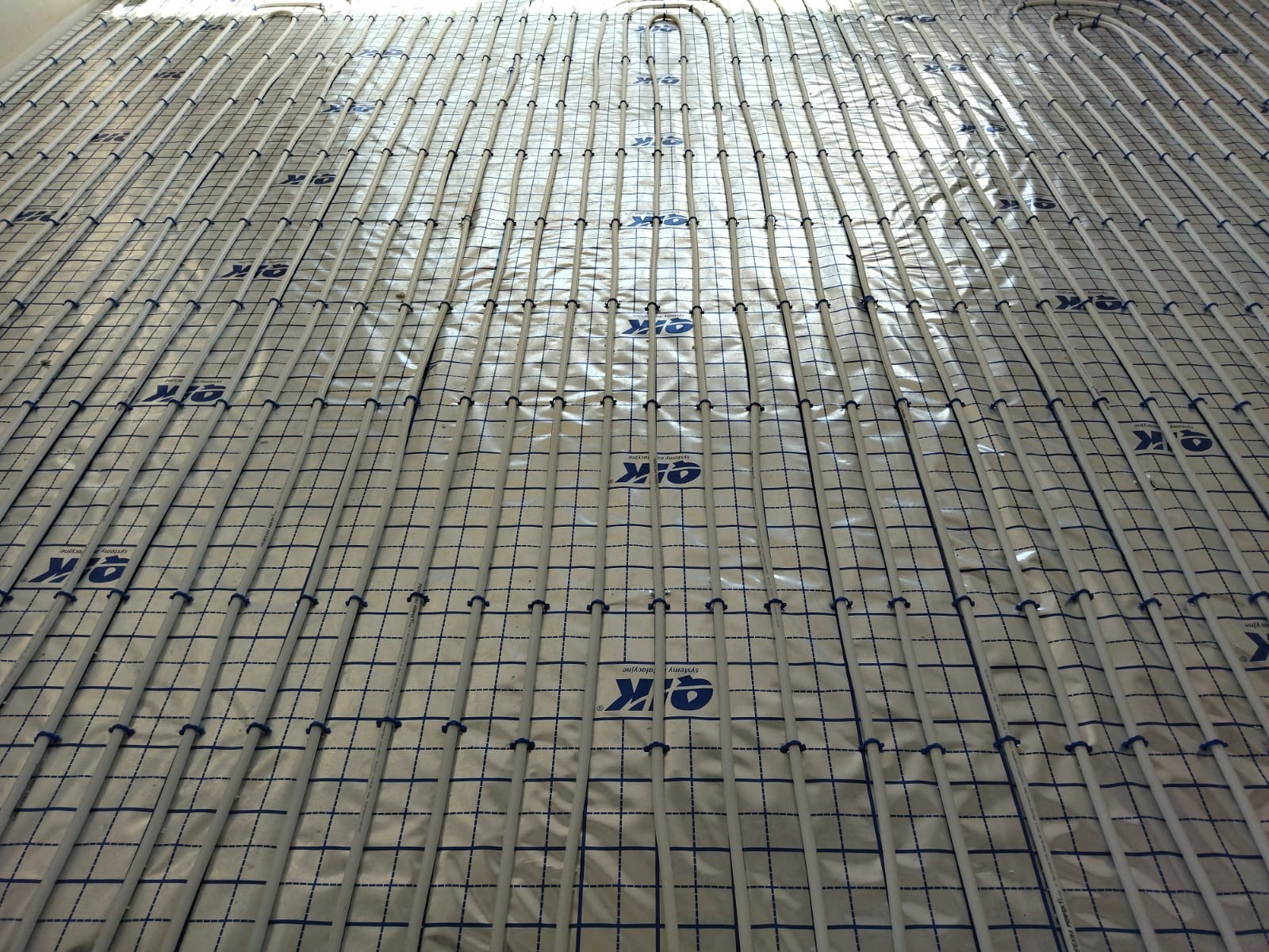Równomiernie rozłożone rury ogrzewania podłogowego na folii z nadrukowaną siatką i logo, przygotowane do zalania wylewką.
