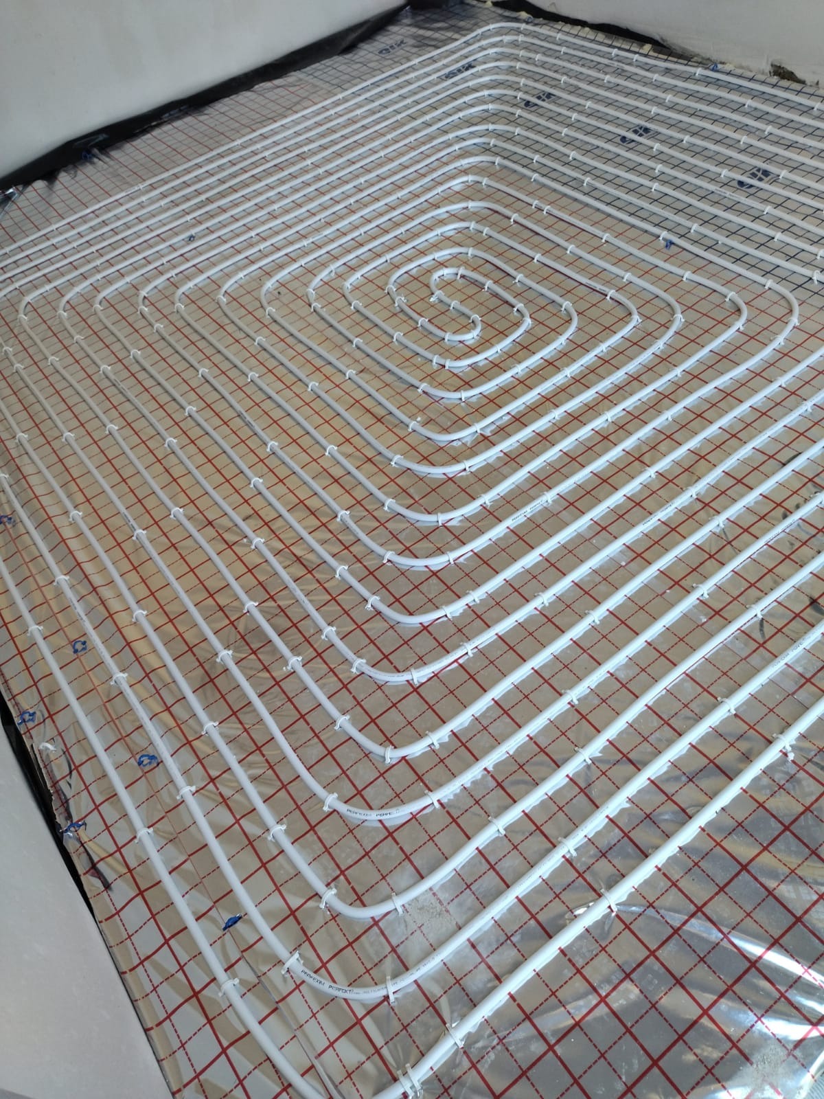 Białe rury ogrzewania podłogowego ułożone spiralnie na folii z czerwoną siatką, widoczne niebieskie klipsy mocujące.