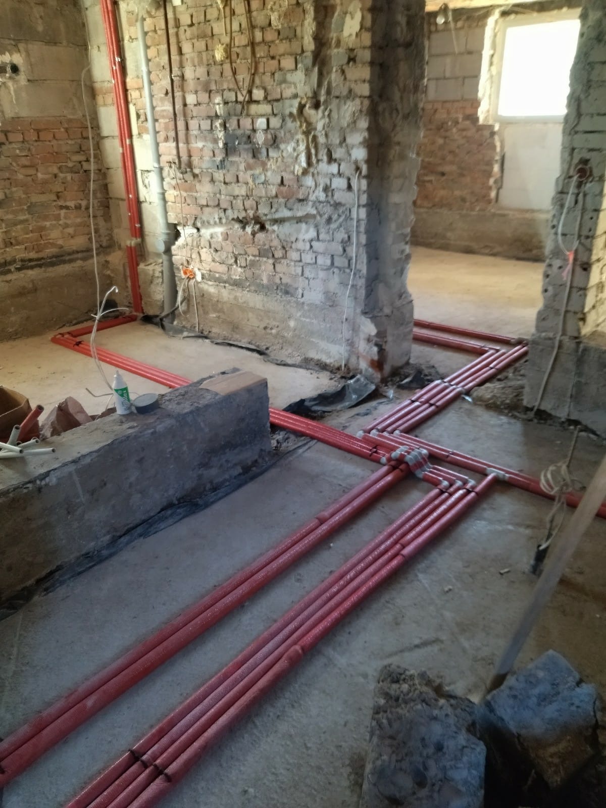 Czerwone rury instalacji grzewczej ułożone na surowym betonie w trakcie remontu, widoczne fragmenty ceglanych ścian i okno w tle.