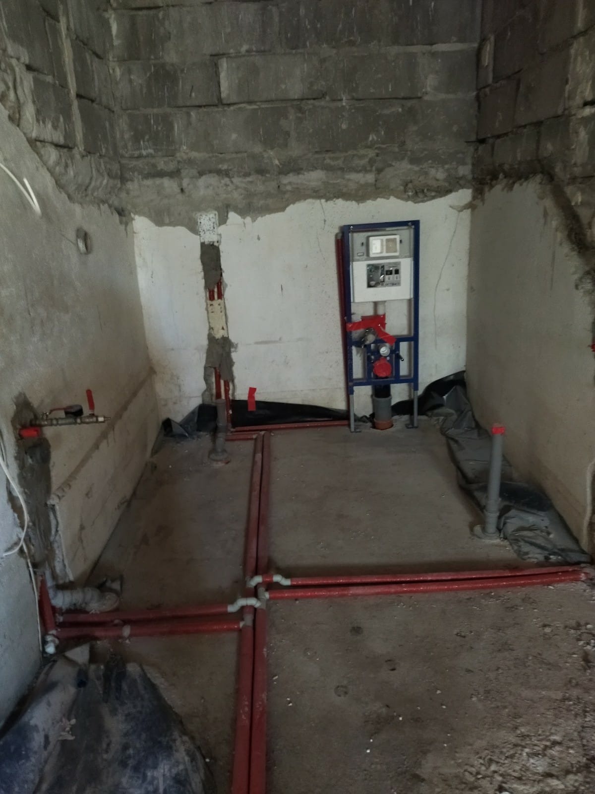 Surowy stan łazienki w trakcie instalacji ogrzewania podłogowego i podtynkowej instalacji WC, widoczne czerwone rury grzewcze na podłodze i stelaż podtynkowy na ścianie.