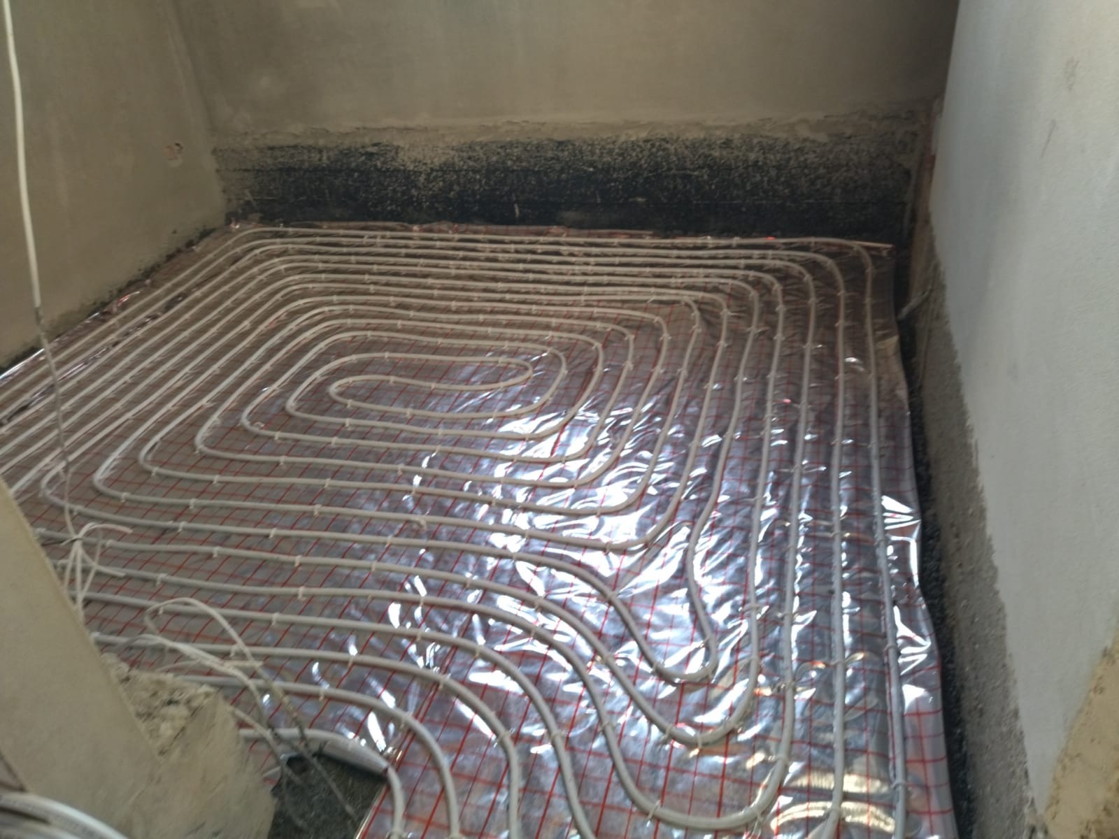 Białe rurki ogrzewania podłogowego ułożone na srebrnej folii z czerwoną siatką w surowym pomieszczeniu przed wylewką.